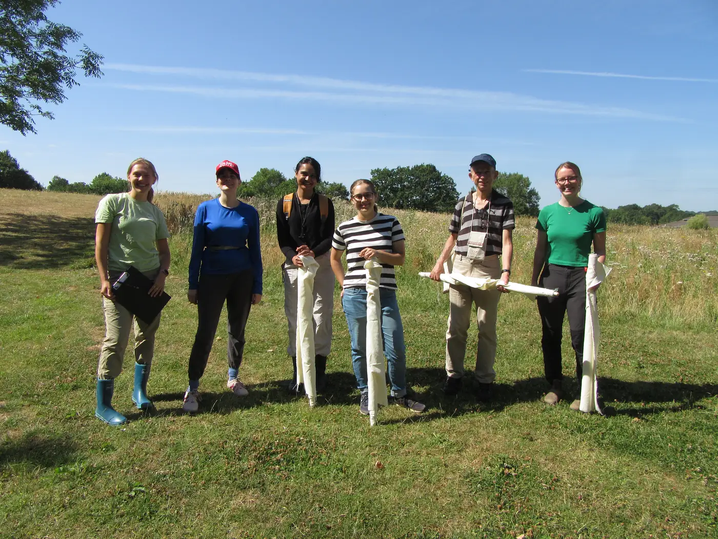 Six people standing in a mown meadow holding rolled-up white tick drag cloths